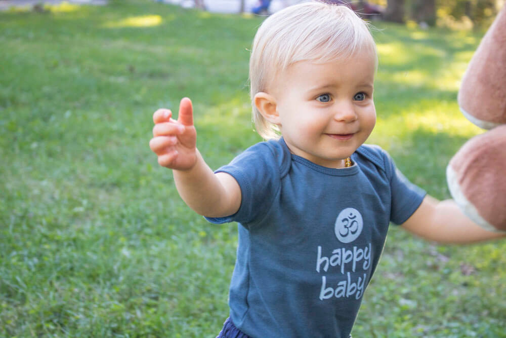 A smiling toddler with blonde hair wearing a blue Natural Baby Clothes happy baby shirt stands on grass, reaching out with both arms. Soul Flower Blog: Hippie Style & Kind Living