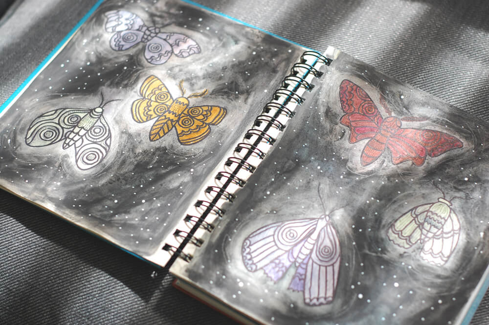 A sketchbook with boho-inspired drawings of six moths on two pages, each outlined in white against a dark, starry background; some moths are colored for an earthy feel, while others remain as line art. Soul Flower Blog: Hippie Style & Kind Living