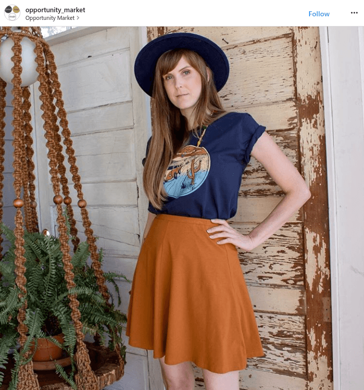 Woman in blue hat, graphic tee, and orange skirt stands by a rustic door and hanging plant, capturing the free-spirited vibe of the Soul Flower Blog.