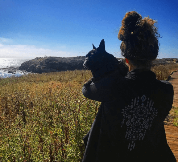 A kind person with hair in a bun holds a small black dog while standing on a wooden boardwalk overlooking the ocean and grassy landscape. Soul Flower Blog: Hippie Style & Kind Living