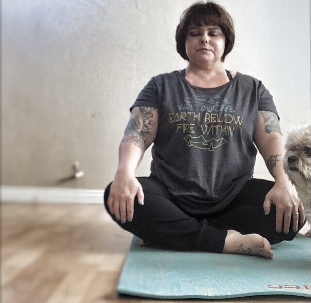 A woman with a boho vibe sits cross-legged on a yoga mat indoors, eyes closed and hands resting on her knees. She wears a t-shirt and black pants. Soul Flower Blog: Hippie Style & Kind Living