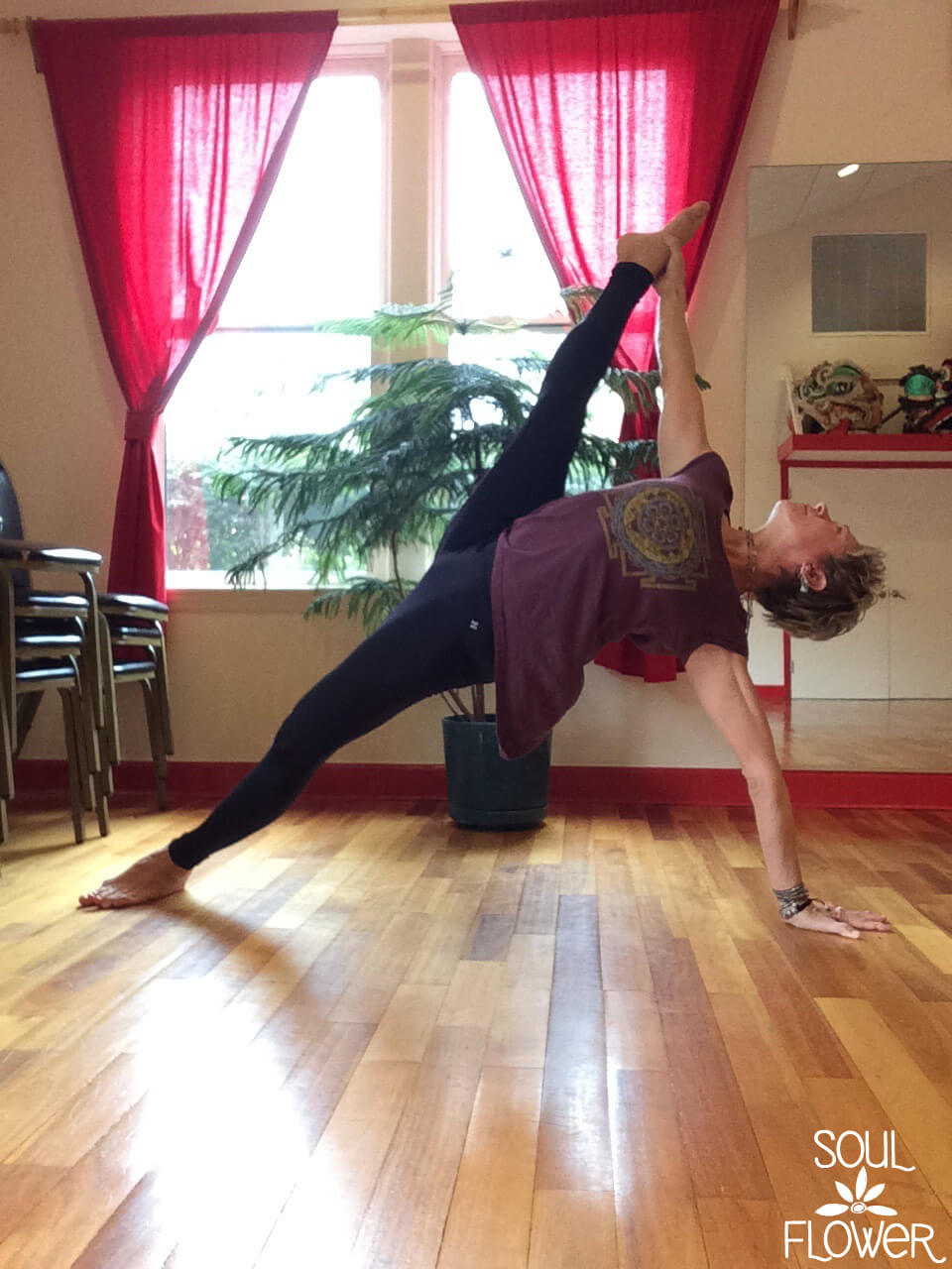 Person performing a side plank yoga pose, balancing on one hand and foot, in a sunlit room with wooden floor, red curtains, and a plant nearby. Soul Flower Blog: Hippie Style & Kind Living