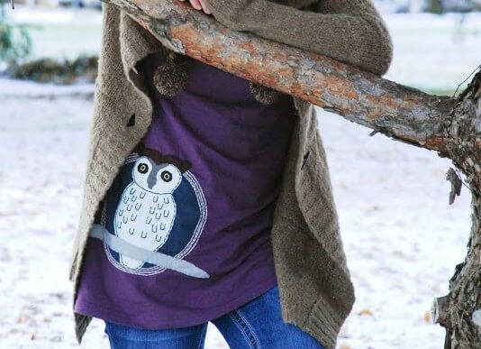 Person wearing a brown sweater and purple shirt with a Woot! Snowy Owl Organic Tunic graphic stands outside, partially obscured by a tree branch. Soul Flower Blog: Hippie Style & Kind Living