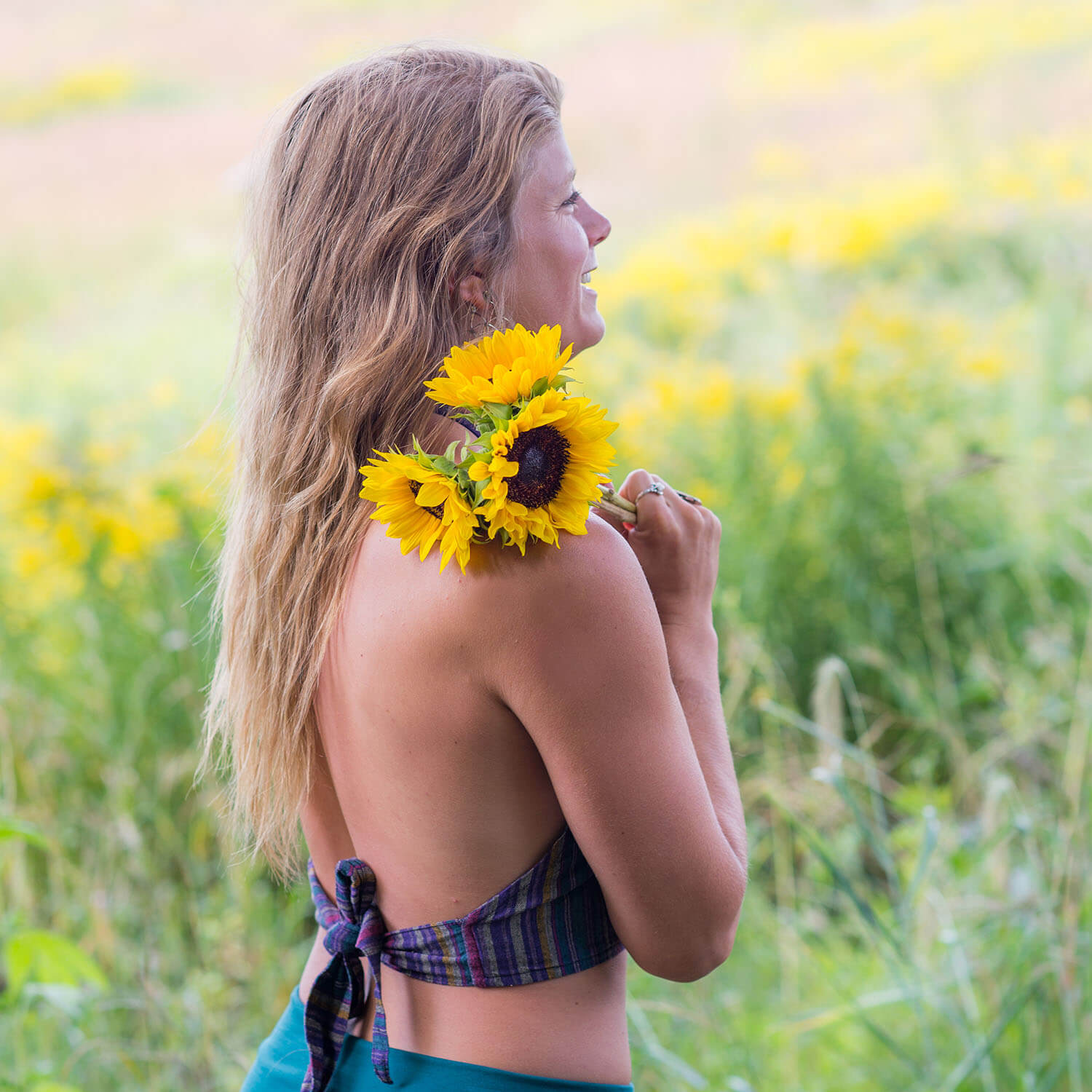 Woman with long hair holds sunflowers on her shoulder, standing in a field of yellow flowers, smiling—radiating the joyful spirit often found on the Soul Flower Blog.