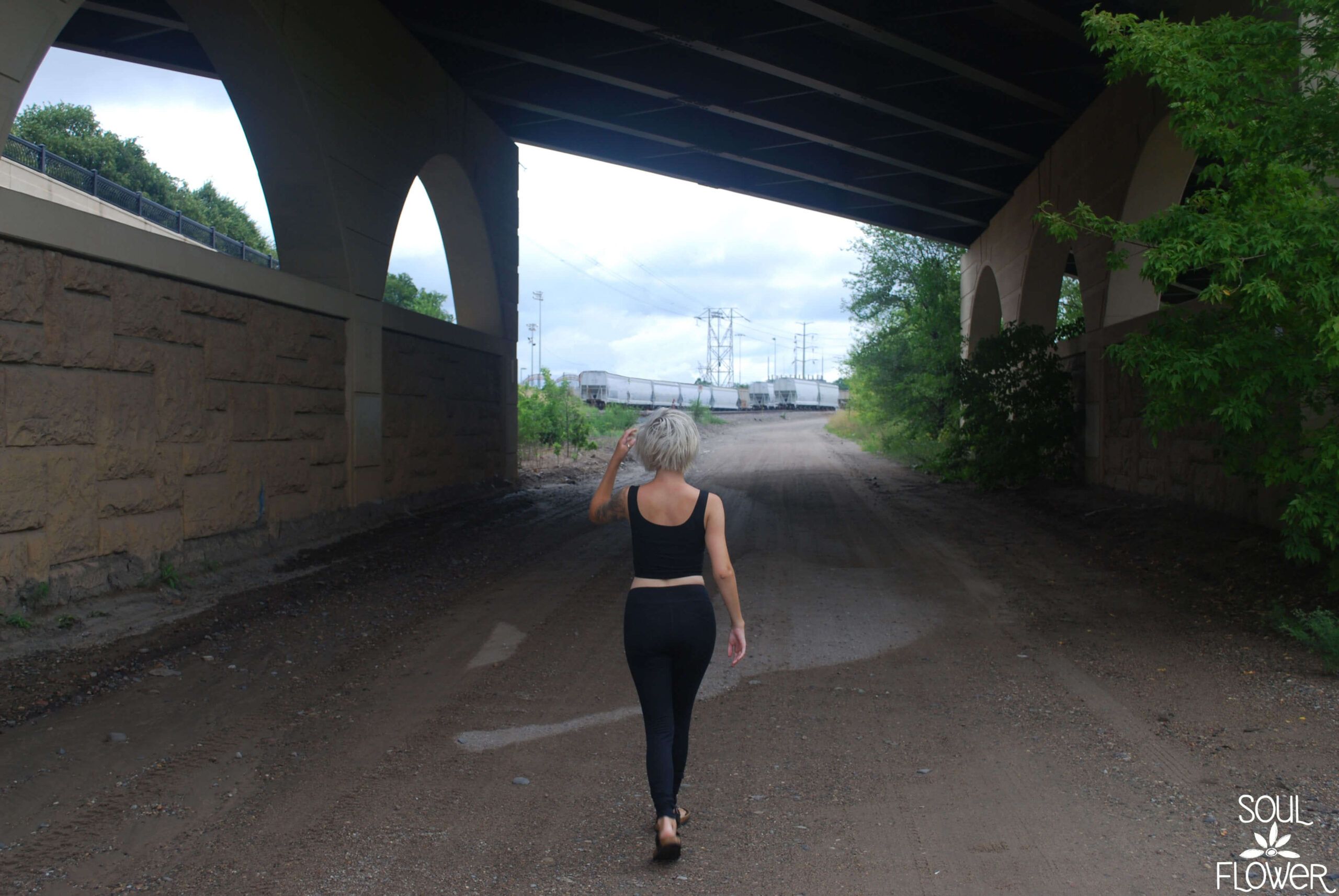 A person with short, light hair walks alone under a large bridge on a dirt path, train cars in the distance. Beneath the overcast sky, it's a moment to jump—and grow your wings on the way down. Soul Flower Blog: Hippie