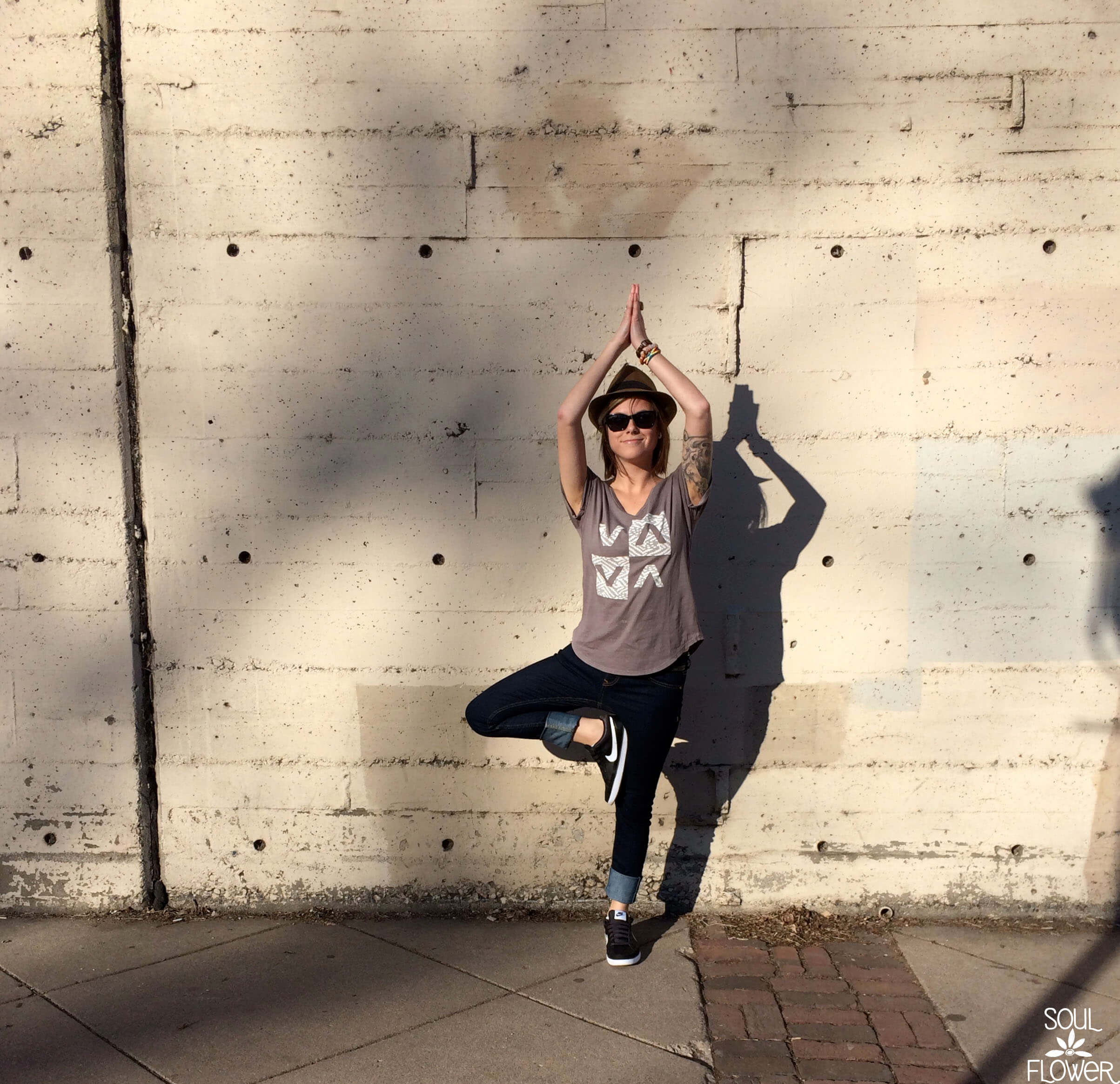 A person wearing sunglasses and a t-shirt stands in a yoga tree pose against a concrete wall, radiating an earthy, hippie vibe, with their shadow visible behind them. Soul Flower Blog: Hippie Style & Kind Living