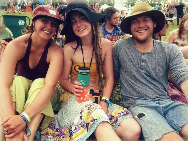 Three young adults sit on grass at an outdoor event, wearing earthy, casual clothes and wide-brimmed hats, smiling at the camera. Other people and tents are visible in the background. Soul Flower Blog: Hippie Style &