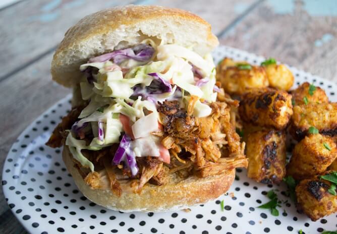 An earthy pulled pork sandwich topped with coleslaw on a bun, served with tater tots on a white plate with black dots—perfect for a hippie-inspired, eco-friendly meal. Soul Flower Blog: Hippie Style & Kind Living