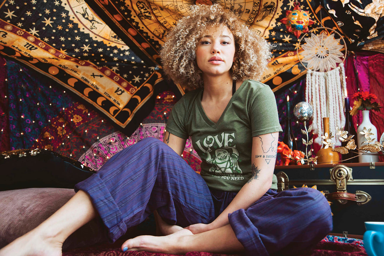 A person with curly hair sits cross-legged on a bed in a colorful, bohemian-style room decorated with tapestries and various ornaments, perhaps pondering ideas from a Hippie Name Generator.