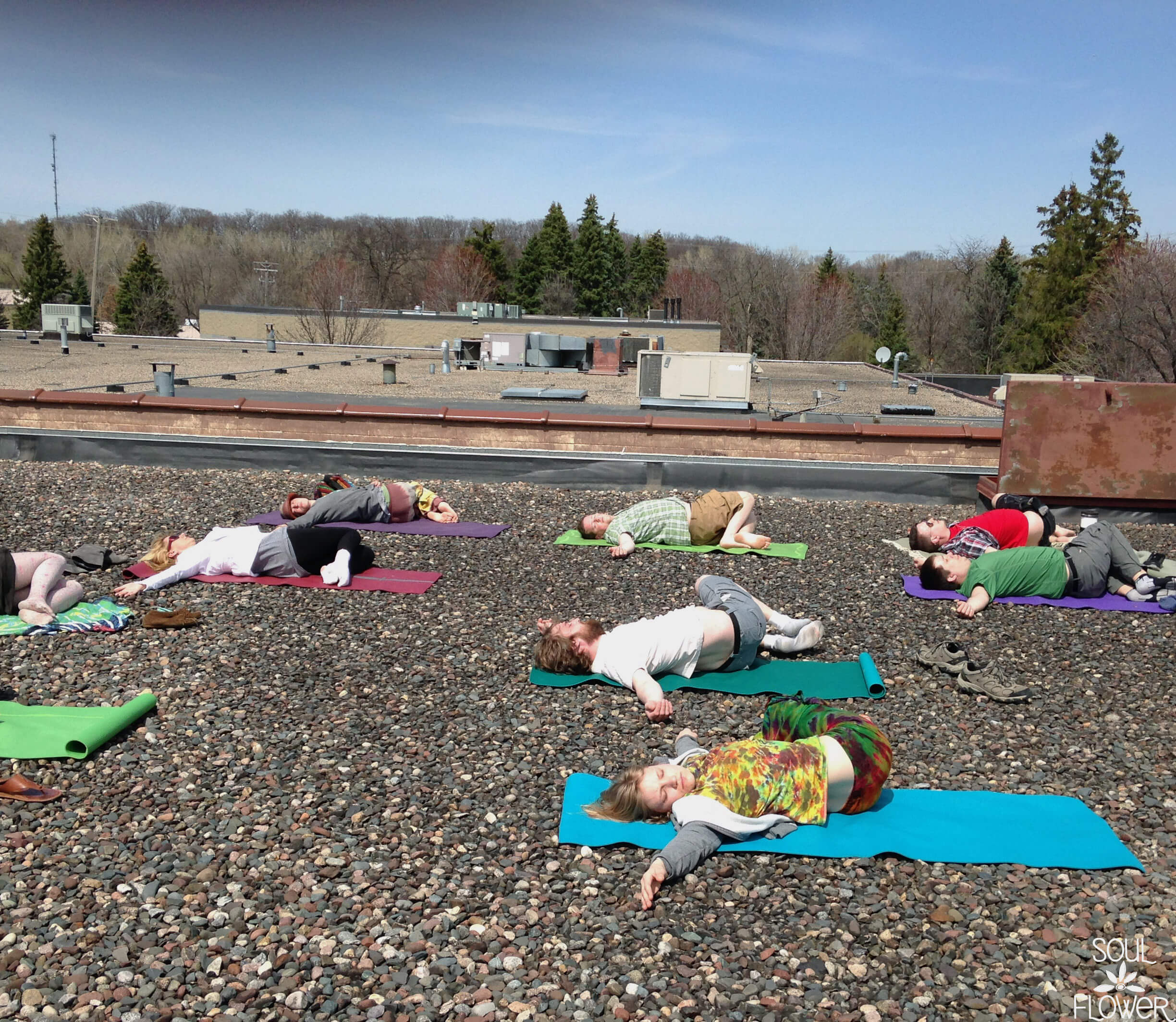 A group of people lying on yoga mats, practicing relaxation or meditation on an earthy rooftop covered with small stones, under a clear sky. Soul Flower Blog: Hippie Style & Kind Living