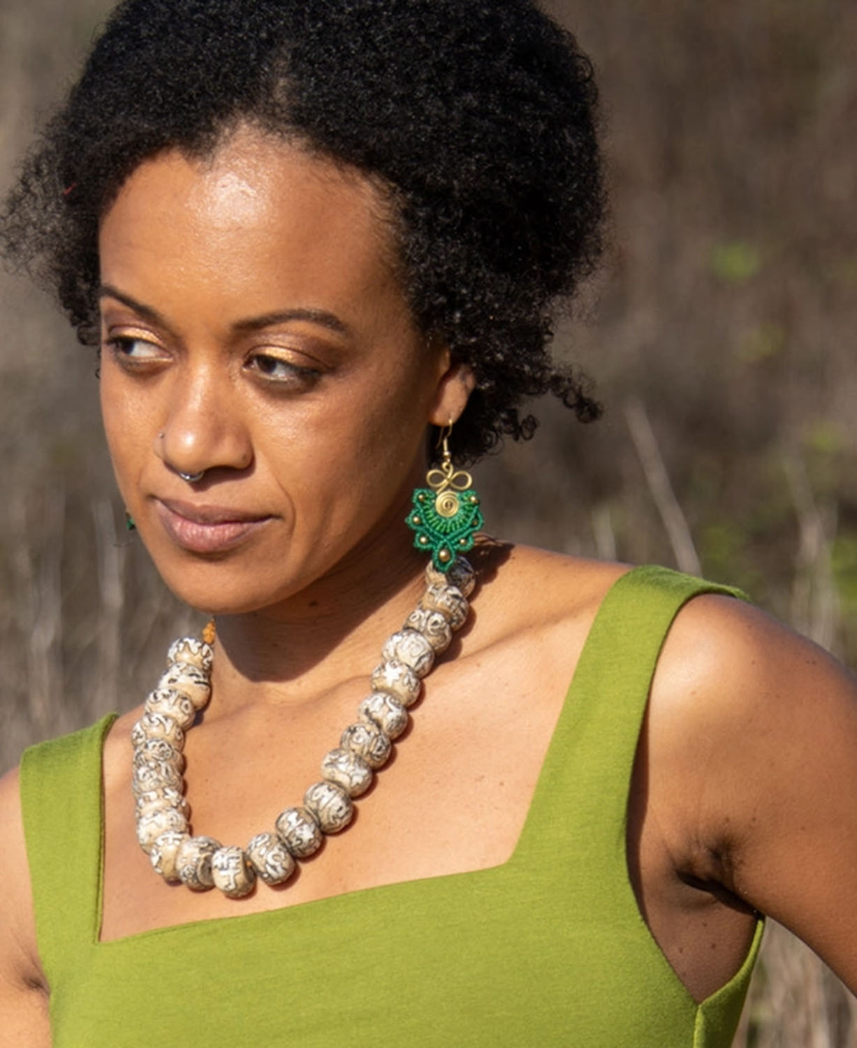 A woman wearing  green and gold dangle earring, photographed outdoors against a blurred natural background.